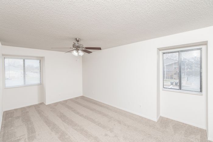 Carpeted bedroom with ceiling fan, two windows with blinds, and neutral walls.