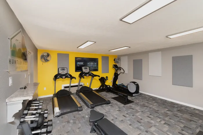 Fitness center at Huntley Ridge Apartments with two treadmills, rowing machine, wall-mounted TV, and bright yellow accent wall.