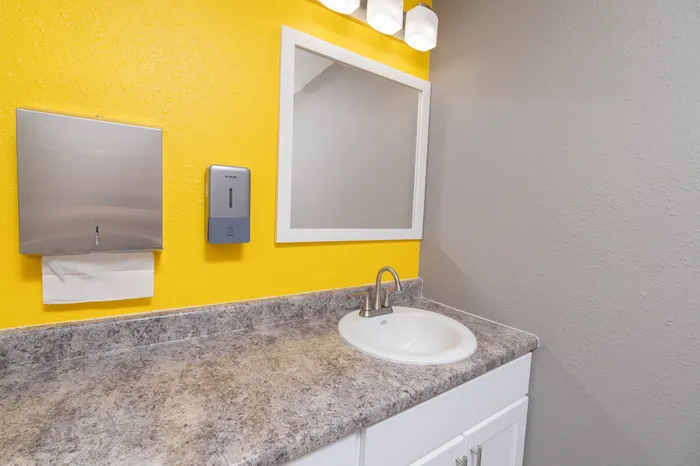 Fitness center sink area with granite-look countertop, white cabinetry, mirror, paper towel dispenser, and yellow accent wall.