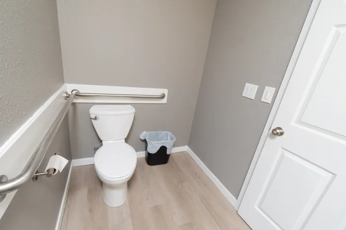 Accessible restroom in fitness center with grab bars, white toilet, light wood flooring, and neutral gray walls.