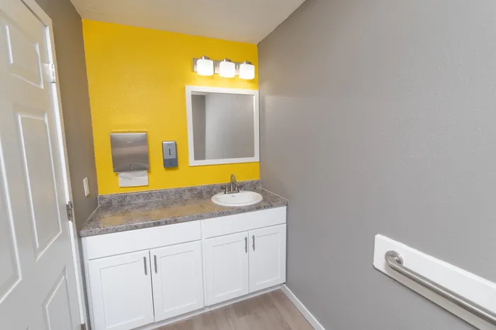 Fitness center restroom vanity with white cabinets, granite-look countertop, mirror, and modern lighting on yellow accent wall.