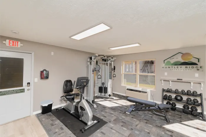 Spacious fitness center at Huntley Ridge Apartments with cardio machines, weight equipment, bench, and natural light.