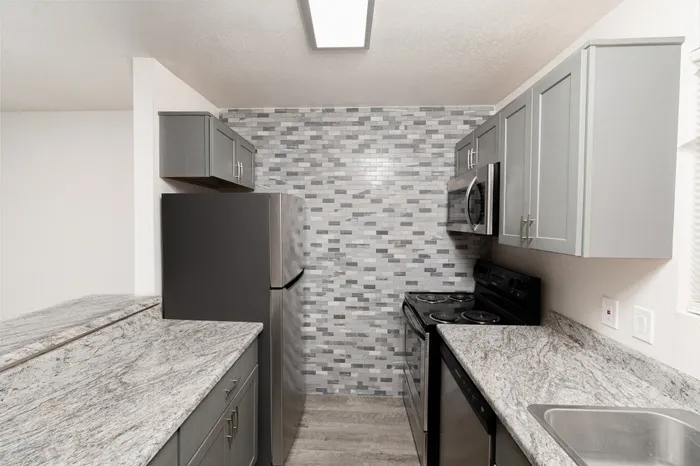 Modern galley kitchen at Hillcrest 90 Apartments with gray cabinets, granite-style countertops, stainless appliances, tile backsplash, and wood-look flooring.