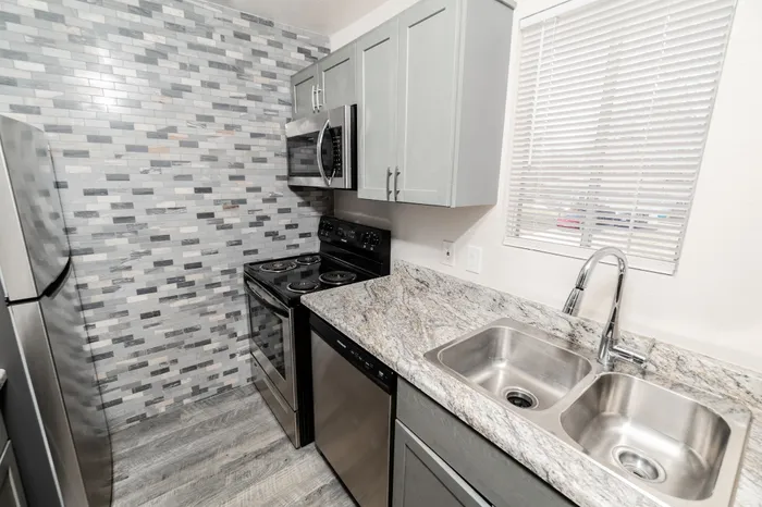 Updated Hillcrest 90 kitchen with double-basin stainless sink, gray cabinets, microwave, electric range, tile backsplash, and window for natural light.