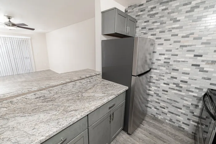 Hillcrest 90 apartment kitchen featuring stainless refrigerator, gray cabinetry, granite-style countertops, tile backsplash, and breakfast bar opening to living room.