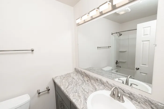 Hillcrest 90 apartment bathroom with granite-style vanity countertop, modern faucet, large mirror with vanity lighting, and tub shower in background.