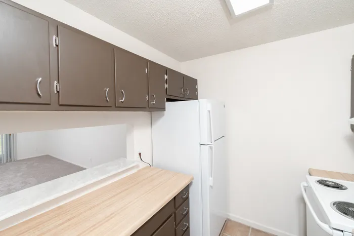 Hillcrest 90 apartment kitchen with brown cabinets, laminate countertops, white refrigerator, and pass-through opening to the living area.