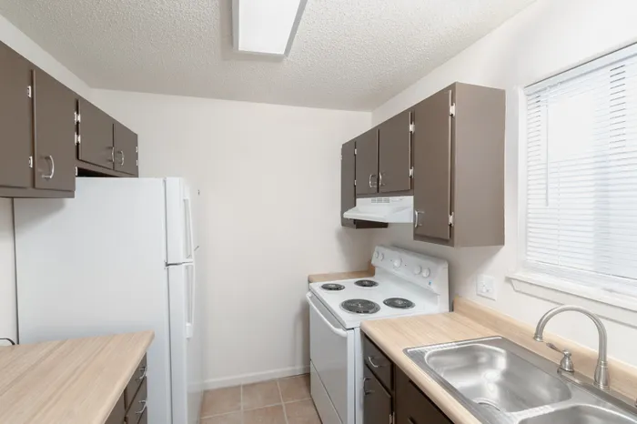 Hillcrest 90 kitchen with brown cabinets, laminate countertops, white appliances, stainless sink, and overhead lighting.