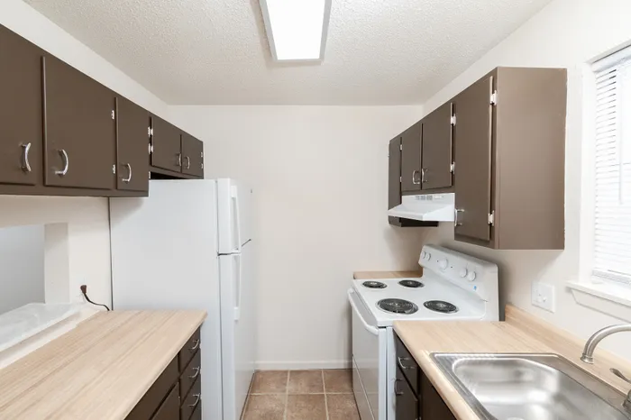 Galley kitchen at Hillcrest 90 Apartments featuring brown cabinetry, white electric stove, refrigerator, stainless sink, and window with blinds.