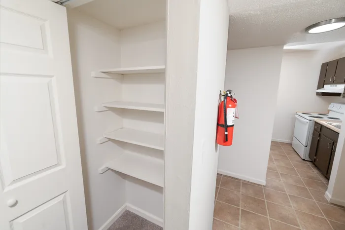 Apartment hallway with built-in white shelving for storage next to a closet door, leading toward the kitchen with tile flooring.
