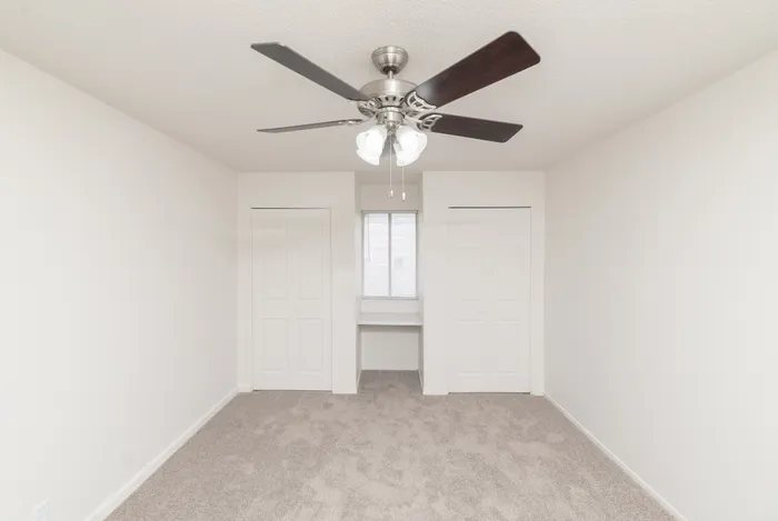 Carpeted bedroom with ceiling fan, window with blinds, and two open closet spaces on either side of a recessed window nook.