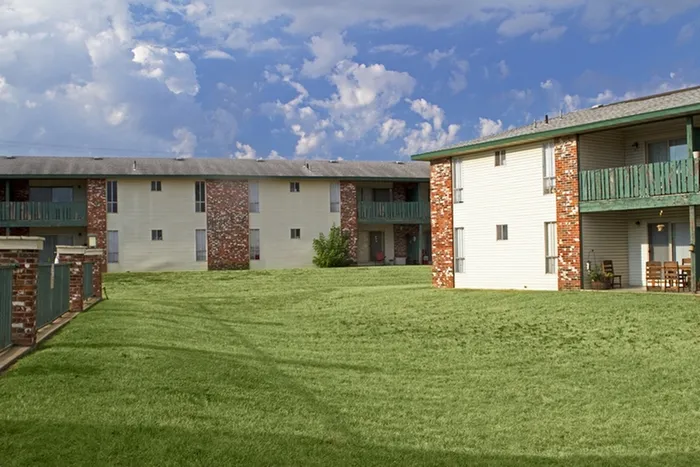 Charming apartment complex with spacious green lawns, brick accents, and inviting balconies under a vibrant blue sky.