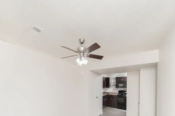 Bright living room with ceiling fan, wood-style flooring, white walls, and open entry leading into the adjacent kitchen area.