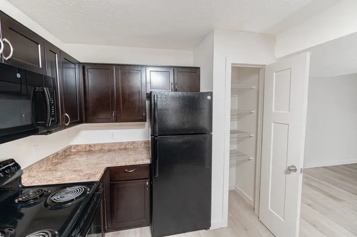 Apartment kitchen with dark wood cabinets, laminate countertops, black appliances, electric range, microwave, refrigerator, and an open pantry closet near the living area.