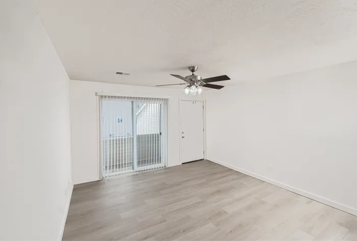 Open living room with light wood-style flooring, white walls, ceiling fan with light, and sliding glass doors with vertical blinds leading to a private patio.