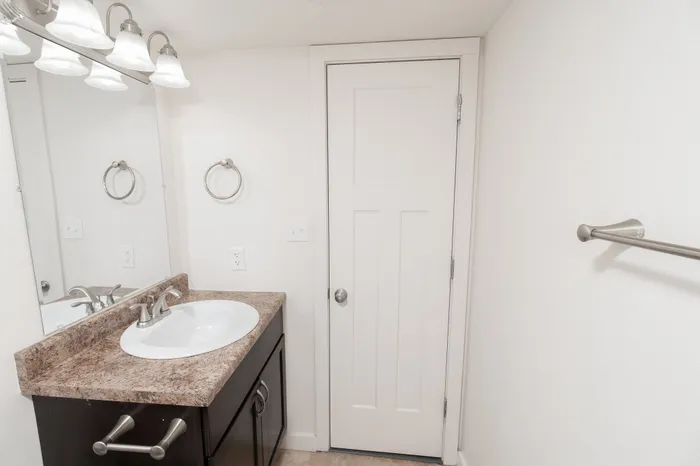 Bathroom vanity with laminate countertop, sink, mirror, overhead lighting, and towel ring next to the bathroom door.