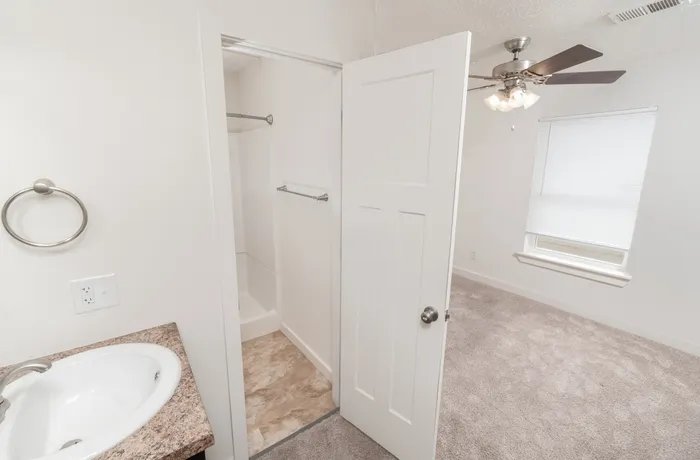 Bathroom with laminate vanity and shower area leading into adjacent bedroom with carpet, ceiling fan, and window with blinds.