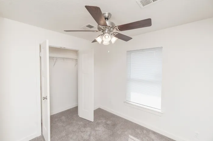 Bedroom with carpet flooring, ceiling fan with light, window with blinds for natural light, and open closet with hanging storage.