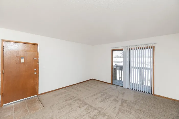 Living room with beige carpet, wood-trim entry door, and sliding glass door to private balcony with vertical blinds.