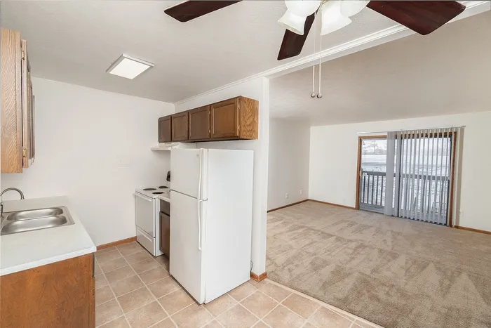 Galley kitchen with wood cabinets, white appliances, and tile flooring, opening into carpeted living room with balcony access.