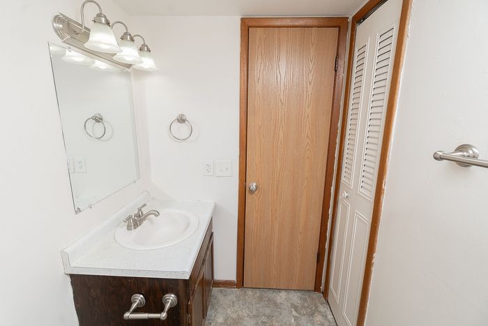 Bathroom vanity with sink, mirror and lighting, wood cabinet storage, and adjacent closet and door.