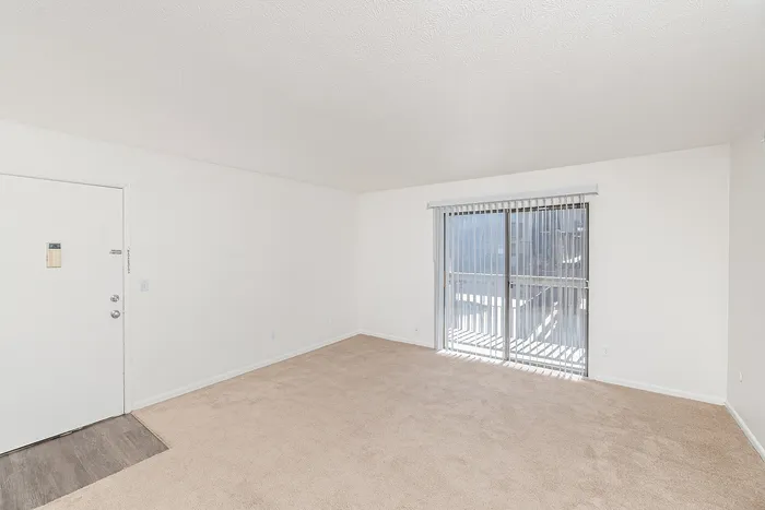 Spacious living room with beige carpet, white walls, and sliding glass door leading to private balcony with vertical blinds and natural light.