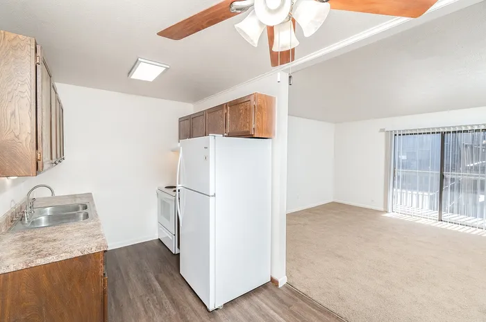 Galley-style kitchen with wood cabinets, laminate countertops, white appliances, and ceiling fan, opening into bright carpeted living room.