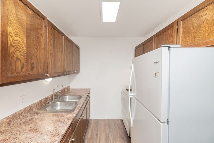 Narrow kitchen with wood cabinetry, double sink, laminate countertops, white refrigerator and stove, and overhead lighting.