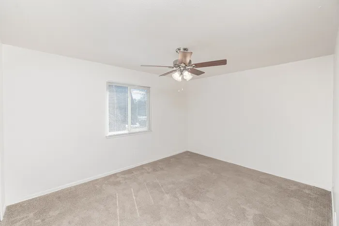 Bright bedroom with neutral carpet, white walls, ceiling fan, and single window with blinds.