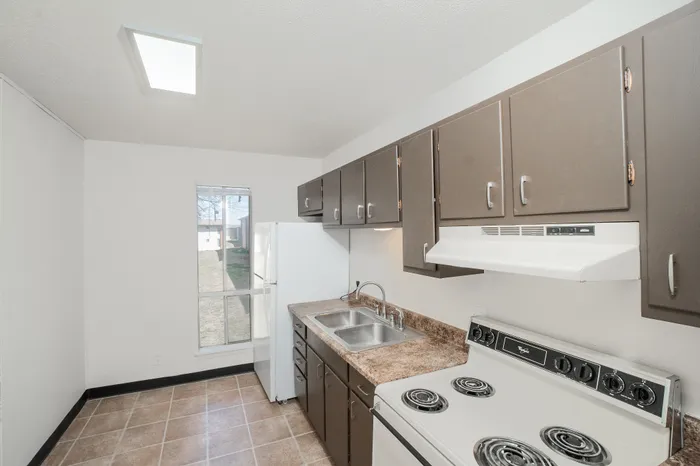 Classic apartment kitchen with brown cabinets, laminate countertops, double sink, electric stove, range hood, and white refrigerator with window lighting