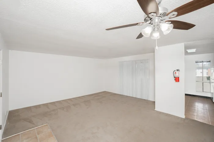 Spacious living room with beige carpet, white walls, ceiling fan with light, and sliding glass door with vertical blinds