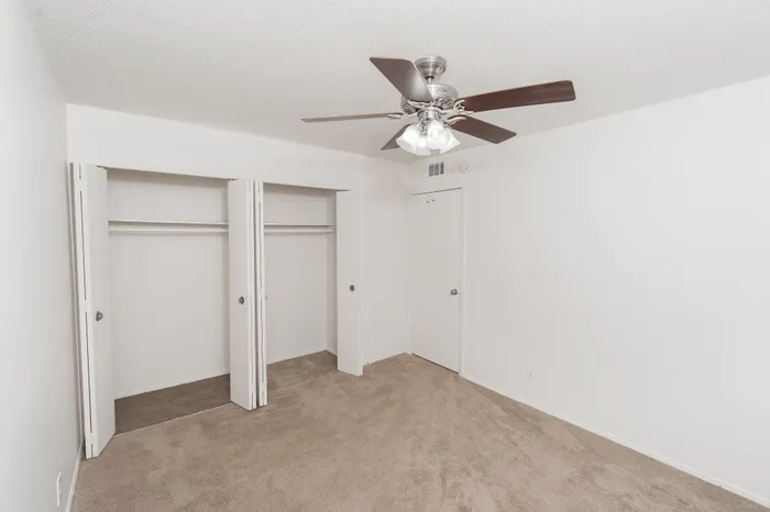 Bedroom with beige carpet, ceiling fan, dual closets with sliding doors, and neutral white walls