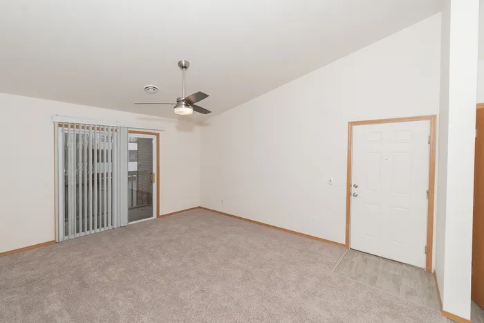 Bright living room with carpet flooring, ceiling fan, large window, and sliding glass door leading to balcony at Plainview Park apartments