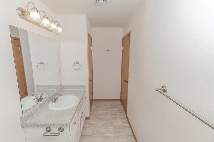 Bathroom vanity area with single sink, large mirror, overhead lighting, storage cabinets, and hallway access in Plainview Park apartment