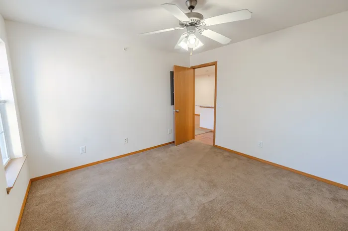 A second angle of the bedroom, showing beige carpet, a ceiling fan, a window with natural light, and an open door leading to the hallway.