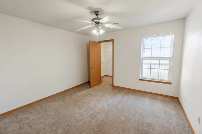 A spacious bedroom with beige carpet, a ceiling fan with lights, a large window with blinds, and an open wooden door leading to a walk-in closet.