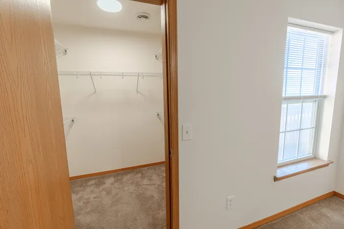 A view of a walk-in closet with wire shelving and neutral-colored walls, adjacent to a window with blinds for natural light.