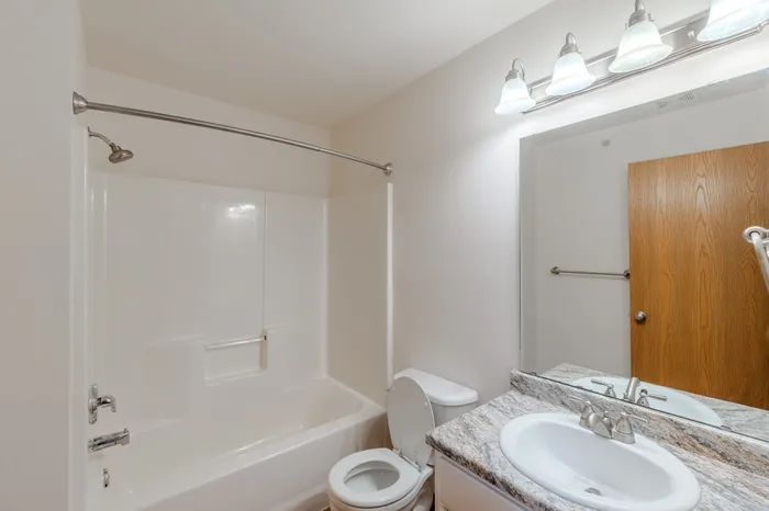 A well-lit bathroom with a white bathtub and shower combination, curved shower rod, granite countertop with a single sink, and a wooden door in the background.