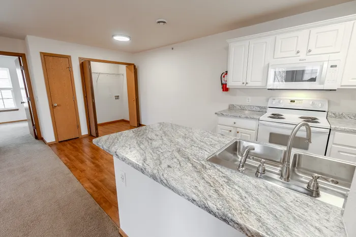 A bright kitchen with white cabinets, granite countertops, a stainless steel sink, and white appliances, including a stove and microwave.