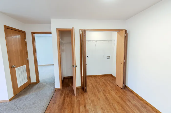 A room with wooden floors featuring open wooden doors leading to a closet with wire shelves, a laundry hookup, and an HVAC vent panel.