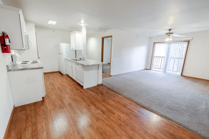 A view from the living room into the open-concept kitchen, featuring wood flooring, carpeted living space, and a sliding glass door to the balcony.