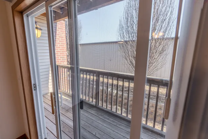 A view through a sliding glass door leading to a wooden balcony, with a bright blue sky and trees visible outside.