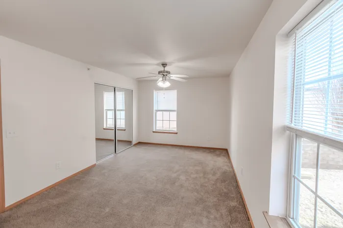 A bedroom with beige carpet, a ceiling fan, two windows, and a mirrored sliding closet door reflecting the interior space.