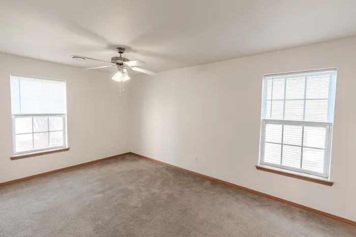 A spacious bedroom with beige carpet, a ceiling fan, and two large windows with white blinds allowing natural light.