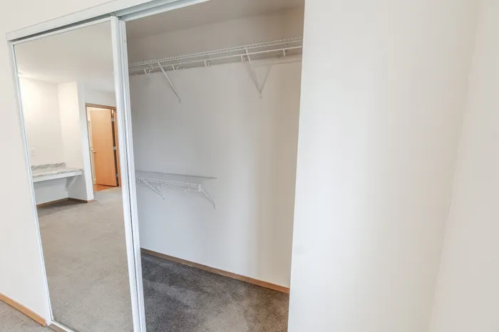 A closet with a sliding mirrored door, featuring wire shelving on the back and side walls. The closet opens into a carpeted bedroom area with a built-in granite vanity visible in the background.