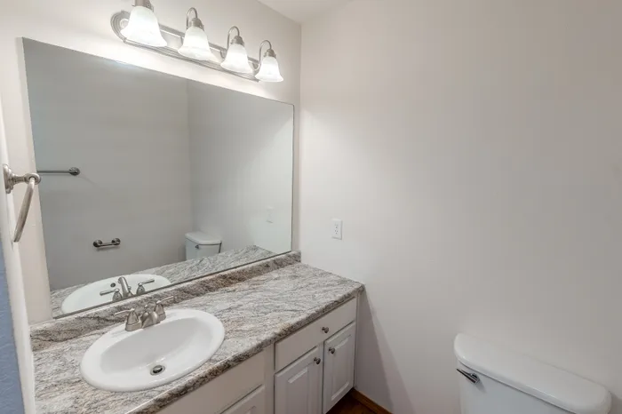 A bathroom featuring a large granite countertop with a single sink, a wide mirror, and a toilet.