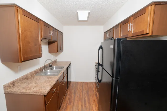 Compact galley kitchen at Rosewood Village Apartments featuring warm wood cabinetry, laminate countertops, double basin sink, black appliances, modern cabinet hardware, and wood style flooring.