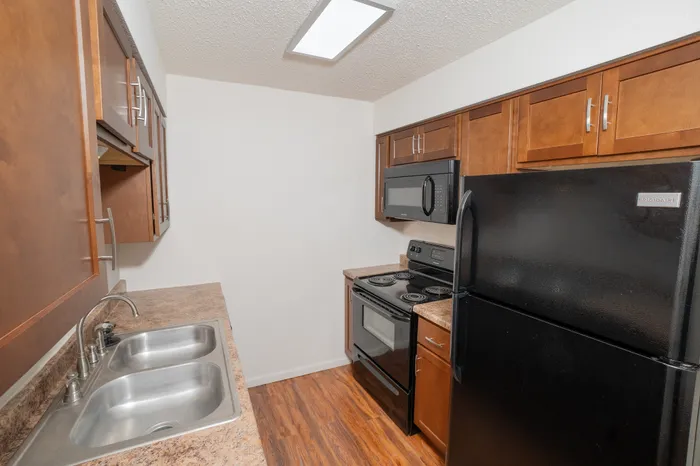 Updated kitchen interior at Rosewood Village Apartments with black refrigerator and range, microwave, wood cabinetry, dual sink, and efficient galley layout with modern finishes.
