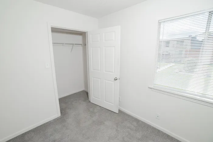 Cozy bedroom interior at Rosewood Village Apartments with plush gray carpeting, bright natural light, white walls, and an open closet with wire shelving for convenient storage.