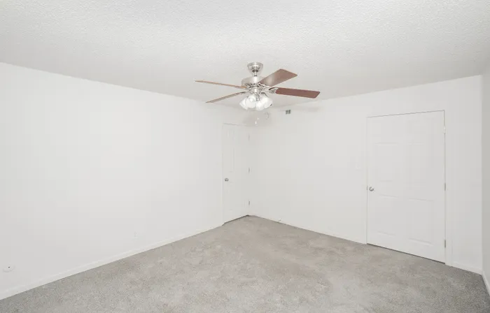 Spacious bedroom at Rosewood Village Apartments featuring soft gray carpet, ceiling fan with lighting, neutral paint tones, and dual closet storage with paneled doors.